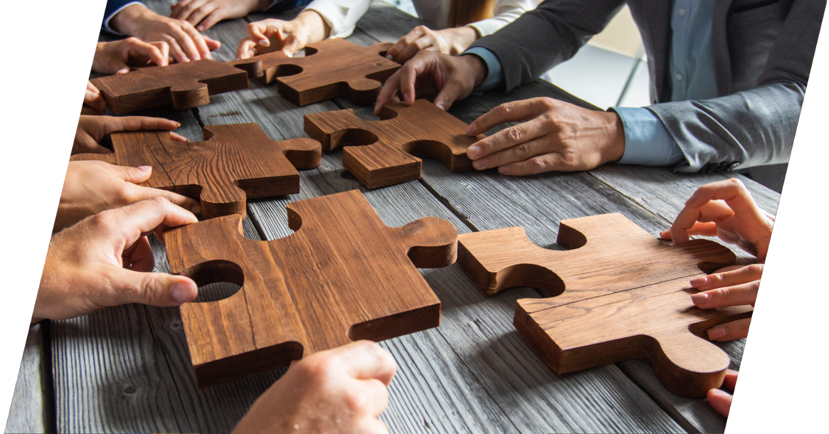 Mehrere Hände halten große Puzzleteile aus Holz in die Mitte einer hölzernen Tischplatte